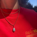 A close-up lifestyle shot of a person wearing a silver teardrop pendant necklace on a beaded chain over a bright red t-shirt with a yellow graphic print.