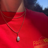 A close-up lifestyle shot of a person wearing a silver teardrop pendant necklace on a beaded chain over a bright red t-shirt with a yellow graphic print.