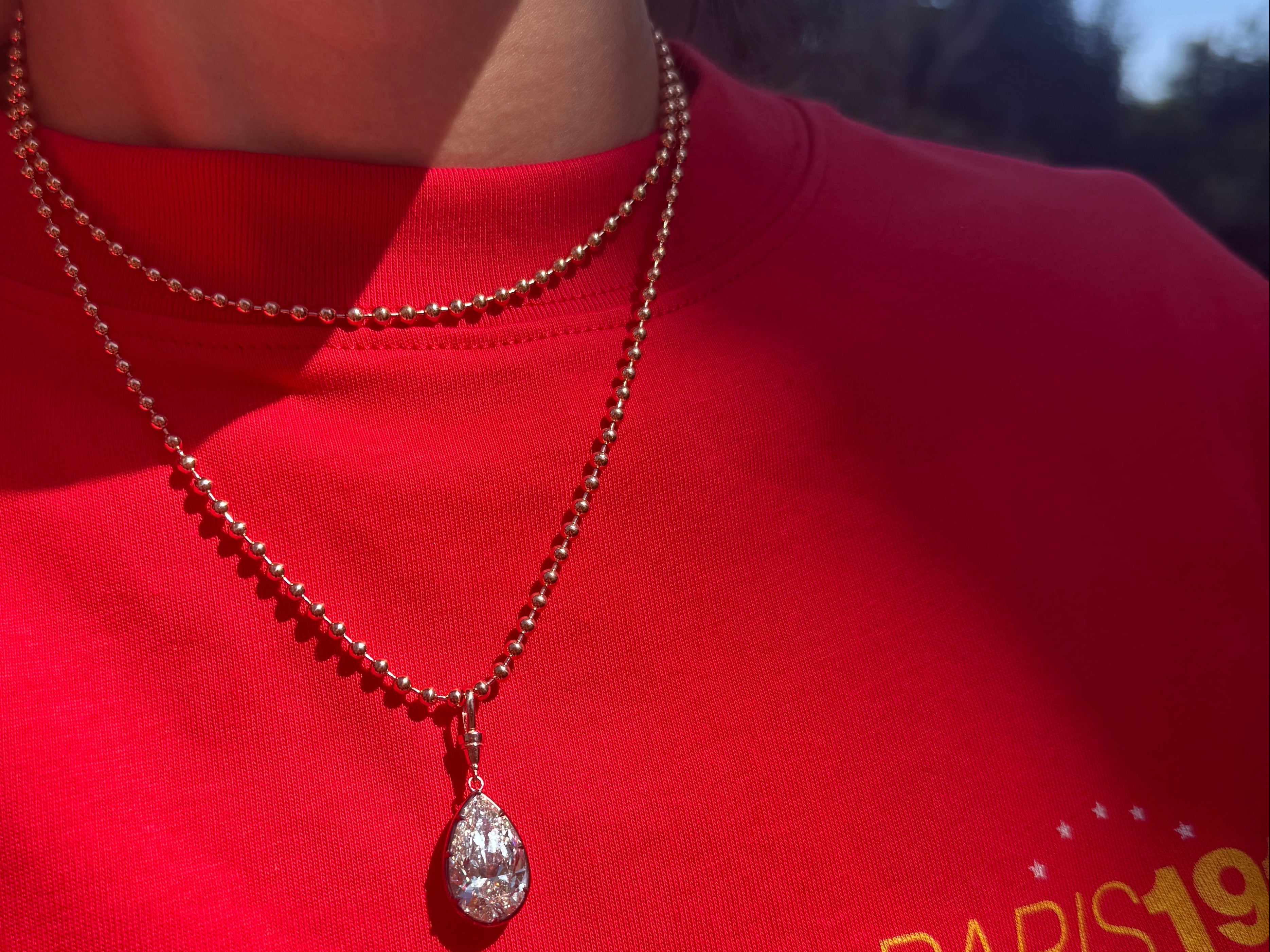 A close-up lifestyle shot of a person wearing a silver teardrop pendant necklace on a beaded chain over a bright red t-shirt with a yellow graphic print.
