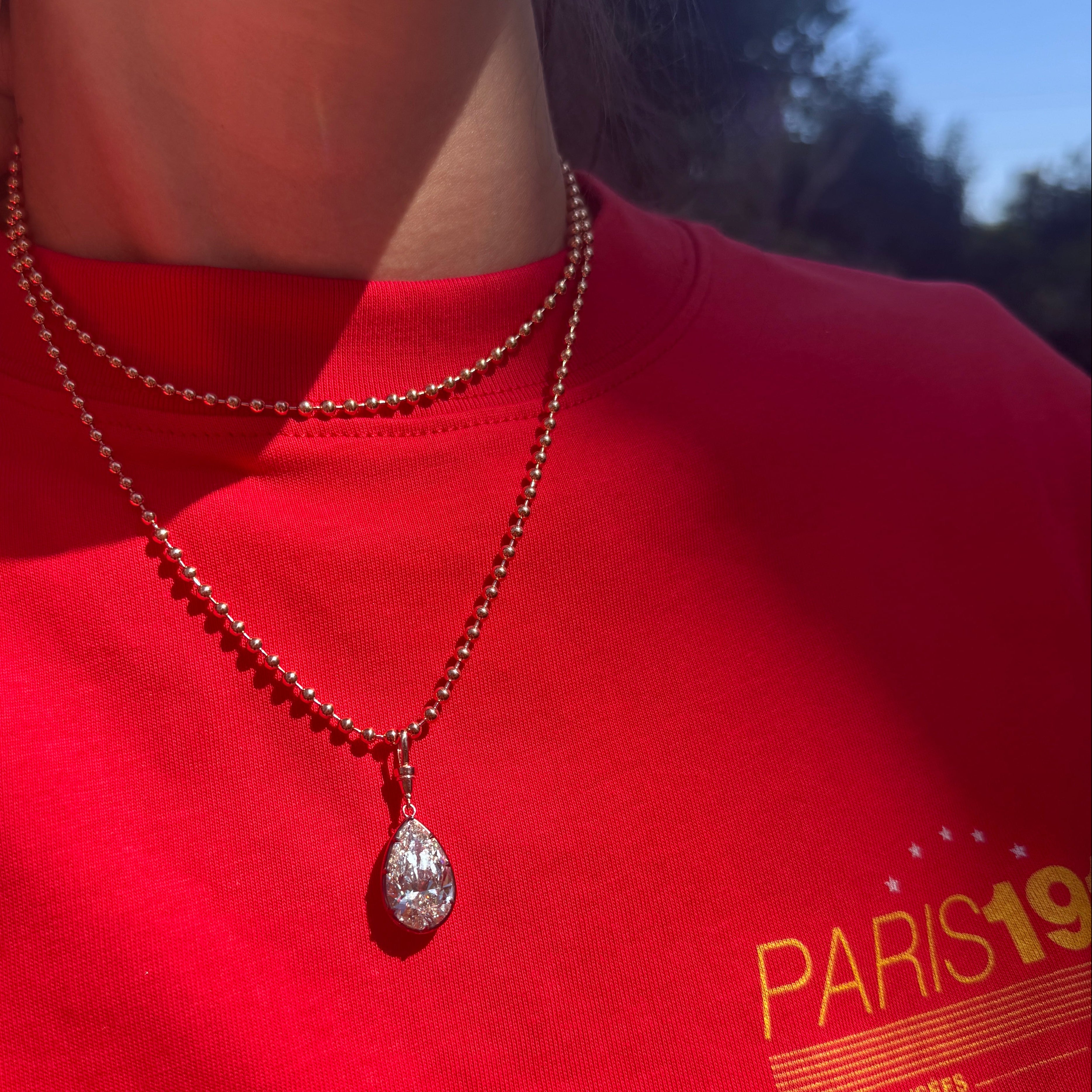 A close-up lifestyle shot of a person wearing a silver teardrop pendant necklace on a beaded chain over a bright red t-shirt with a yellow graphic print.