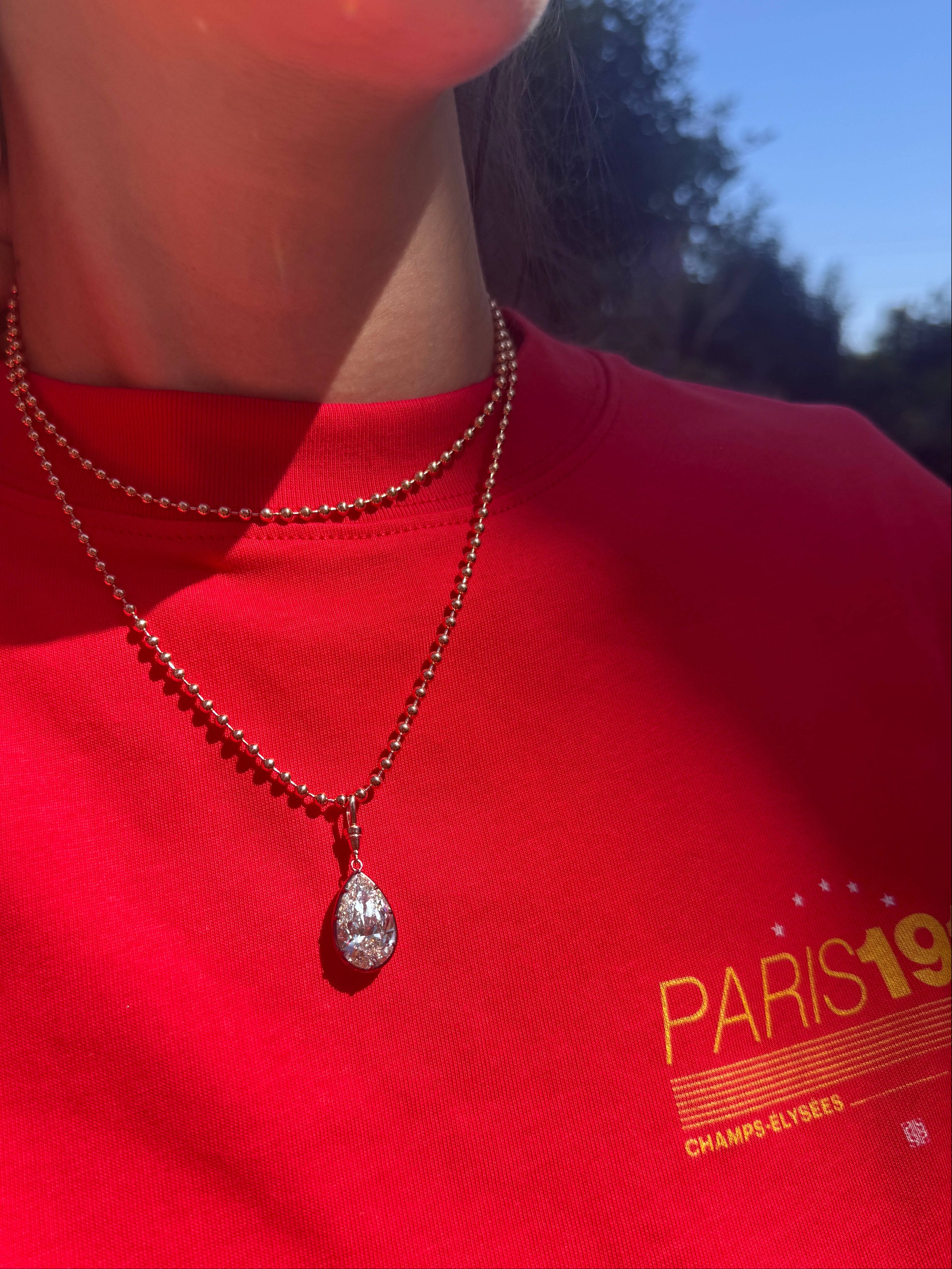 A close-up lifestyle shot of a person wearing a silver teardrop pendant necklace on a beaded chain over a bright red t-shirt with a yellow graphic print.